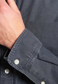 Chemise grise à boutonnage devant en tissu texturé, avec un poignet retroussé, des surpiqûres visibles et des boutons blancs au niveau du poignet.