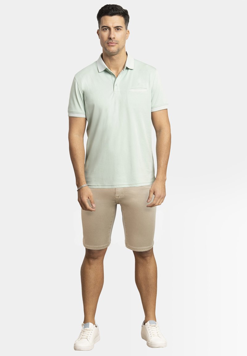 Camisa polo verde claro de manga corta y cuello, combinada con pantalones cortos beiges. El modelo lleva zapatillas blancas y está de pie contra un fondo liso.