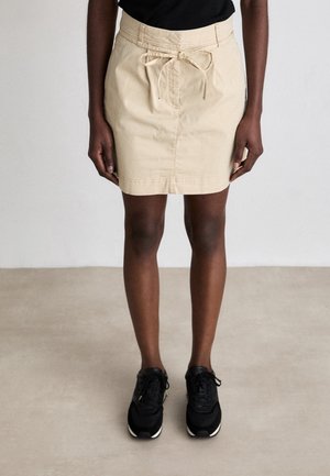 Person wearing a beige knee-length skirt with a front tie belt, black top, and black sneakers standing on a light gray floor.