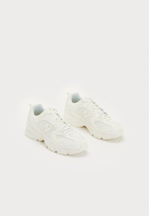 Zapatillas deportivas blancas con malla y cuero liso, que presentan perforaciones sutiles, una puntera redondeada y una suela de goma blanca con detalle de logo.