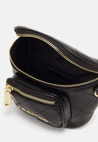 Černá prošívaná kožená kabelka se zlatými taháky zipu, otevřenou hlavní přihrádku a přední kapsou na bílém pozadí.