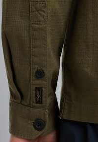 Camicia a maniche lunghe verde oliva con tessuto testurizzato, dotata di bottoni neri e un'etichetta tessuta. I polsini includono dettagli di cucitura e una linguetta.