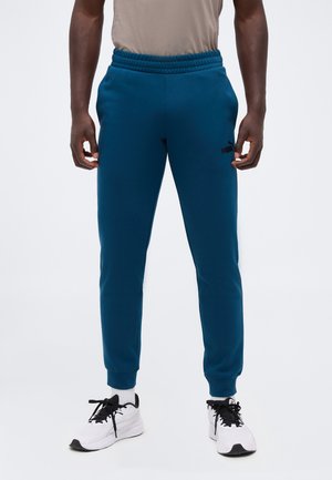 Homme portant un pantalon de survêtement bleu Puma, une chemise beige, des chaussettes blanches et des baskets blanches avec des lacets noirs, debout devant un fond blanc.