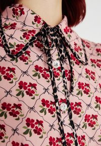 Blusa rosa con motivo floreale che presenta fiori rossi e foglie verdi. Dettagli notevoli includono un colletto leopardato in bianco e nero e bottoni.