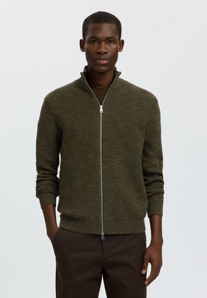 Mann trägt einen olivgrünen Reißverschluss-Pullover und dunkelbraune Hose, steht vor einem schlichten hellen Hintergrund mit den Händen in den Taschen.