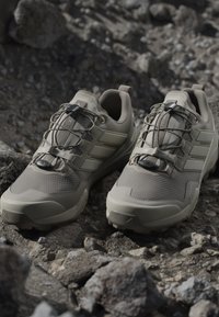 Light beige trail running shoes with textured mesh upper, elastic laces, and cushioned sole, set against a rocky background.