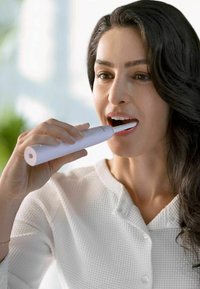 Elektrische tandenborstel met een witte cilindrische behuizing en een borstelkop. Iemand gebruikt de tandenborstel en draagt een gestructureerd wit shirt.