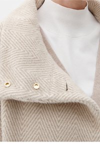 Primo piano di un cappotto in lana a spina di pesce beige stratificato con bottoni dorati sopra un dolcevita bianco a coste e un maglione beige chiaro.