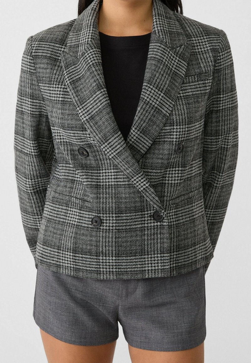 Femme portant un blazer à carreaux gris à double boutonnage sur un top noir et un short gris cintré sur un fond neutre.