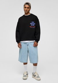 Zwarte sweatshirt met blauwe en rode opdruk op de zak, gecombineerd met lichtblauwe denim shorts. Witte sokken en meerkleurige sneakers maken de look compleet.