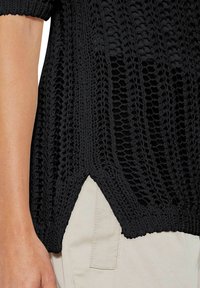 Schwarzes gestricktes Oberteil mit lockerer, offener Webart, gerippten Bündchen und seitlichem Schlitz, darüber gefertigt aus leichtem, glattem Stoff.