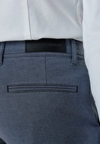 Mörkblå texturerade byxor med ett fiskbensmönster. Har en läderlapp med texten "SHAPING NEW TOMORROW" och en sidoficka.