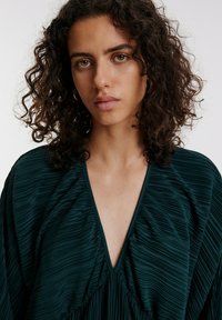 Dunkelgrüne, plissierte Bluse mit tiefem V-Ausschnitt und weiten Ärmeln, aus strukturiertem Stoff und minimalistischem Design. Weiches, leichtes Material.