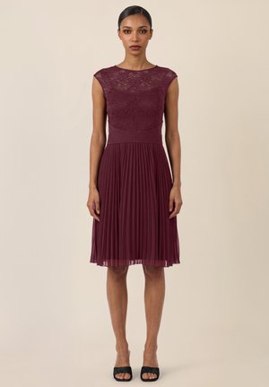 Frau in einem ärmellosen, knielangen bordeauxroten Kleid mit Spitzenoberteil und plisziertem Chiffonrock, stehend vor einem einfarbigen beigen Hintergrund.