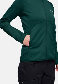 Giacca zip-up verde scuro con una finitura testurizzata, dotata di tasca con zip e design aderente. Abbinata a pantaloni cargo neri.