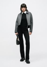 Žena nosiaca sivú skrátenú bundu cez čierny sveter a nohavice, drží čiernu kabelku, stojaca pred jednoduchým bielym pozadím.