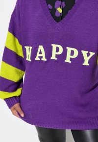Fioletowy sweter z dzianiny z dekoltem w szpic, z żółtymi paskami na rękawach i słowem "HAPPY" w wyraźnych żółtych literach na przodzie.