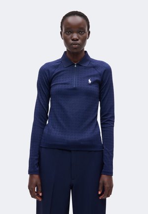 Femme portant un polo bleu marine à manches longues avec une fermeture éclair et un petit logo blanc sur la poitrine, associé à un pantalon foncé.