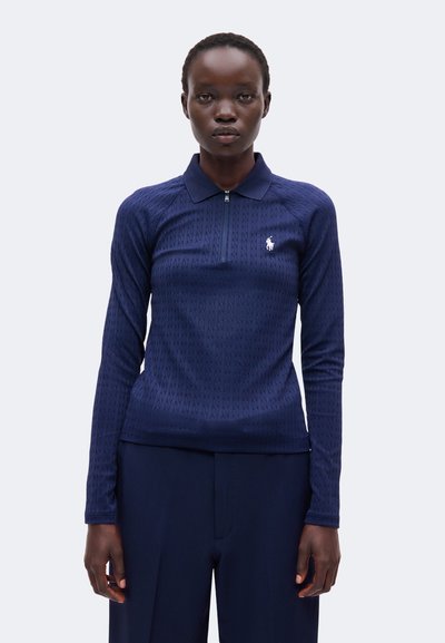 Femme portant un polo bleu marine à manches longues avec une fermeture éclair et un petit logo blanc sur la poitrine, associé à un pantalon foncé.