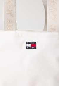 Balta rankinė su lygiu paviršiumi, kurią puošia audinio logotipo užklotas mėlynos, raudonos ir baltos spalvų. Dirželiai turi audinį primenančią tekstūrą.