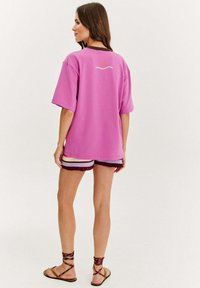 Velká růžová tričko s kulatým výstřihem, krátkými rukávy a malým logem na zadní straně. V kombinaci s barevnými pletenými šortkami a sandály.