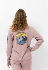 Lange mouwen, zachtroze pullover met een afbeelding van bergen en golven op de achterkant. De tekst luidt: "BERG EN ZEE WAAR IK WIL ZIJN."