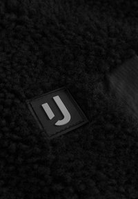 Tessuto in pile nero con superficie testurizzata. Presenta un'etichetta nera quadrata con il logo bianco "UJ" e un accento nero liscio nelle vicinanze.