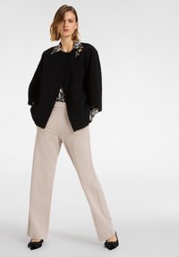 Zwarte, gestructureerde, oversized jas met brede mouwen, gelaagd over een gebloemde blouse, gecombineerd met beige broek en zwarte schoenen versierd met een strik.