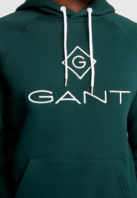 Sudadera verde oscuro hecha de tela suave, con un gran logo blanco de "GANT" y cordones. Incluye un bolsillo tipo canguro en la parte delantera.