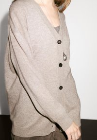 Beige stickad cardigan med V-ringning, fem svarta knappar och långa, lösa ärmar. Lättviktig textur; bärs över en grå topp.