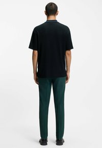Polo shirt nero a maniche corte con design testurizzato, abbinato a pantaloni verdi su misura e scarpe nere, visto da dietro.