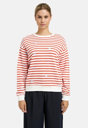 Woman wearing a white sweater with horizontal red stripes and small blue heart patterns, paired with loose black pants.