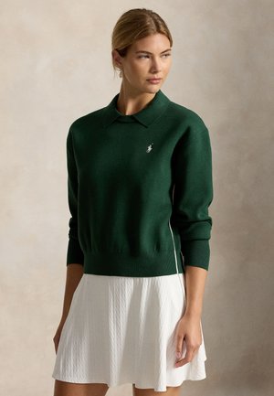 Femme portant un pull vert foncé à col avec un petit logo et une jupe blanche texturée, debout devant un fond neutre.