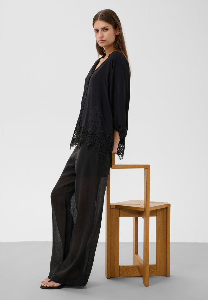 Jeune femme en blouse noire ample avec détails en dentelle et pantalon noir large, appuyée sur une chaise en bois devant un fond uni.