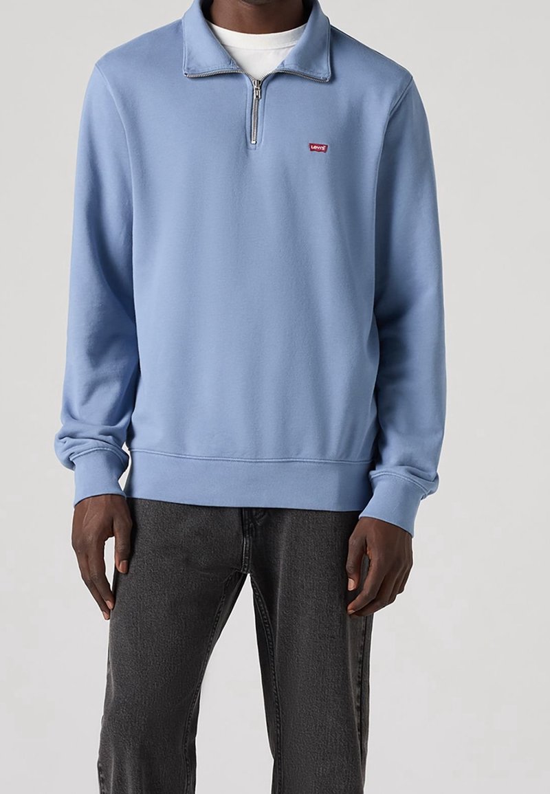 Ljusblå sweatshirt med half-zip krage, ribbad nederkant och ärmslut. Har en liten röd logotyp på bröstet, tillverkad av mjukt material.