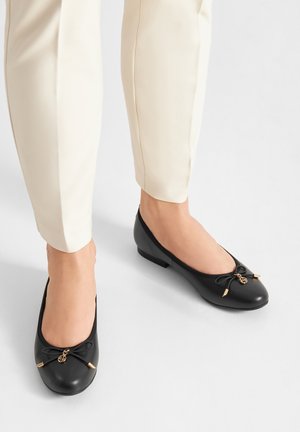 Černé baleríny z kůže se zaoblenou špičkou, ozdobnou mašlí a zlatými kovovými doplňky. Nízký podpatek, hladký povrch a jednoduchý design.