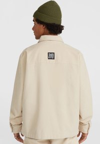 Beige corduroy overshirt met lange mouwen, voorzien van een logo patch op de achterkant en knoopsluitingen aan de mouwen, gedragen met een groene geribbelde beanie.