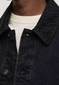 Chaqueta de denim negra con cuello puntiagudo, costuras visibles como detalles y dos botones delanteros. Debajo se ve una camisa de color crema.
