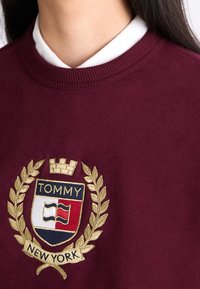 Sudadera burdeos con cuello de canalé, que presenta un emblema bordado con una corona, hojas de laurel y el texto "TOMMY NEW YORK".