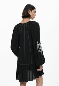 Schwarzes Kleid mit langen Puffärmeln, einem gestuften Rock, vertikalen weißen Nähten und einem gestickten floralen Design am linken Ärmel.