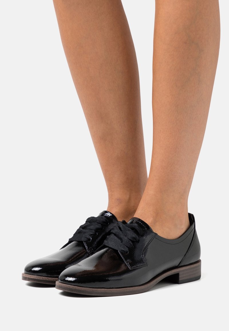 Chaussures Oxford en cuir noir brillant avec de larges lacets noirs, portées pieds nus, et talons empilés bas de couleur marron.