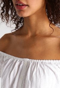 Blusa blanca sin hombros con cuello volado, detalle de botones en la parte delantera y una tela texturizada con sutiles líneas verticales.