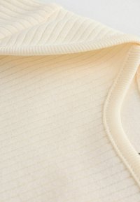 Rippstoff-Oberteil in hellem Beige, mit weicher Textur und rundem Ausschnitt, versehen mit sichtbaren Nähdetails entlang der Kanten.