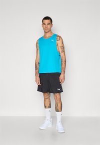 Tirkizna bezrukavna sportska majica s bijelim logom, u kombinaciji s crnim šortsem i bijelim sportskim cipelama, stoji na jednostavnoj pozadini.
