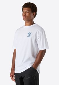 Unge mand med kort, krøllet hår iført en hvid t-shirt med et lille blåt tegneserie-logo og sorte bukser, stående med hænderne i lommerne.