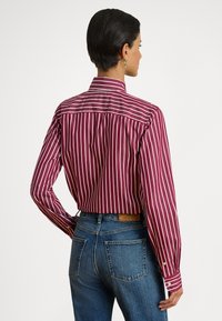 Κόκκινο και λευκό ριγέ πουκάμισο με μακριά μανίκια και ελαφρώς εφαρμοστό πίσω μέρος, σε συνδυασμό με μπλε τζιν τζιν. Διαθέτει κλασικό γιακά και μανσέτες με κουμπιά.