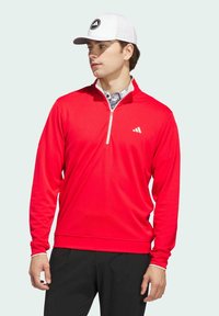 Red long-sleeve pullover with a quarter-zip collar, featuring a white logo on the chest and a textured fabric; paired with a white cap.