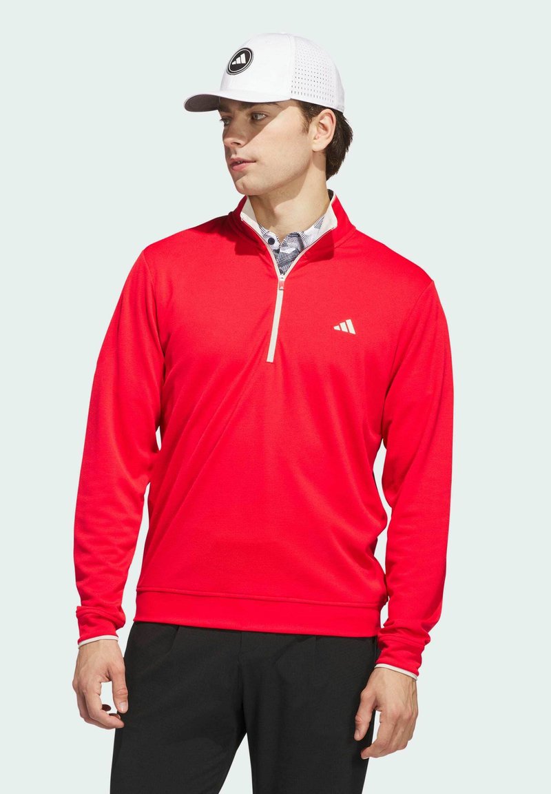 Red long-sleeve pullover with a quarter-zip collar, featuring a white logo on the chest and a textured fabric; paired with a white cap.
