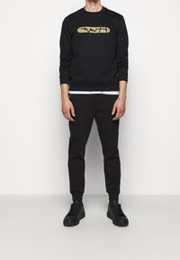 Felpa nera con grafica dorata, abbinata a pantaloni jogger neri. Presenta scollo tondo, maniche lunghe e polsini alla caviglia. Indossata con sneaker nere.