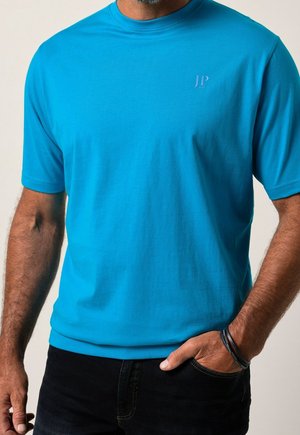 Uomo che indossa una maglietta a maniche corte di un blu brillante con il logo "JP 1880", jeans neri e braccialetto nero intrecciato, mano in tasca.